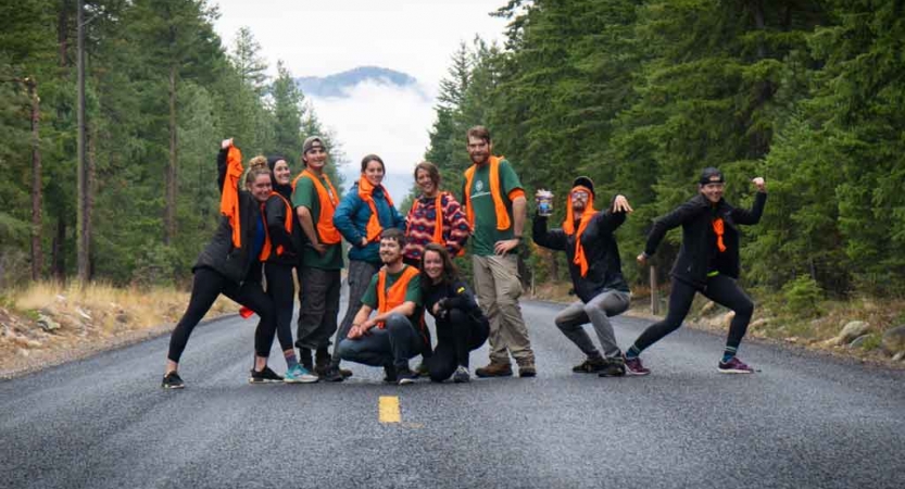 a group of gap year students wearing orange vests pause for a photo on a road between green trees. They have been doing service projects with outward bound. 